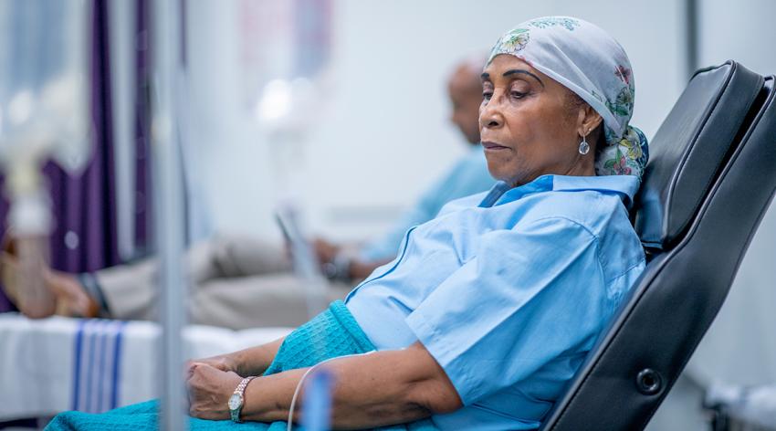 A woman wearing a head scarf lies on a hospital bed and looks to the side. She is wearing a head scarf and a hospital gown and there is a IV drip next to her.