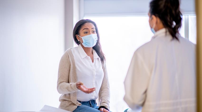 A female patient of African descent is sitting on the examination table during a medical check up with a doctor. They are wearing face masks to prevent the spread of germs.