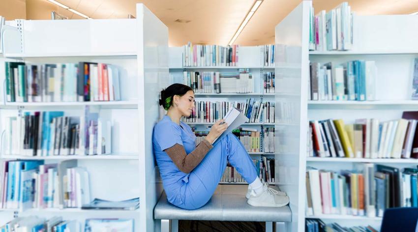 A female student wearing blue scrubs sits on a bookcase reading in a library.