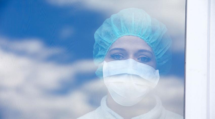 A medical worker in a mask and hairnet stares sadly out of a window